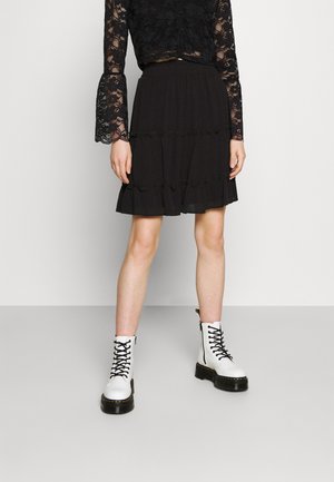 Woman wearing a black lace long-sleeve top, black ruffled skirt, and white lace-up ankle boots standing against a plain background.