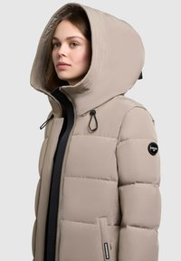 Chaqueta acolchada en beige claro, con una gran capucha, cierre delantero con cremallera y detalles a presión. Incluye un parche con el logo en la manga y bolsillos laterales.