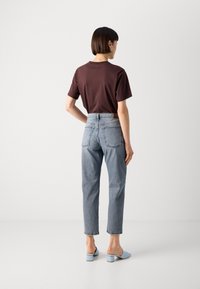 Dunkelbraunes Baumwoll-T-Shirt, hellblaue hochgeschnittene Jeans und pastellblaue Absatz-Pantoletten. Gerade Beine mit sichtbaren Hintertaschen.