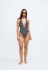 Μαύρο ολόσωμο μαγιό με λευκό κείμενο σε χειρόγραφη γραφή, βαθύ ντεκολτέ σε σχήμα V, λουριά που δένουν γύρω από τον λαιμό και ψηλές ανοίγματα στα πόδια.