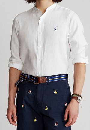 Personne portant une chemise en lin blanche boutonnée avec les manches retroussées, un pantalon bleu marine avec des voiliers brodés, une ceinture rayée et une montre-bracelet avec bracelet.