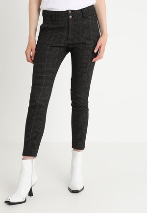 Slim-fit dark plaid trousers with front buttons worn with white heeled ankle boots and a tucked-in white top.