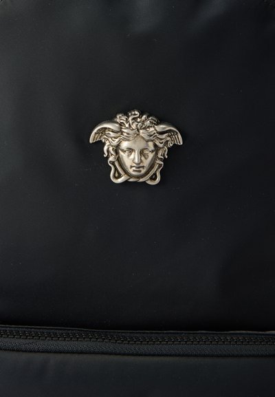 Έμβλημα κεφαλής Μέδουσας από μέταλλο, τοποθετημένο στο κέντρο πάνω σε μαύρο υφασμάτινο φόντο, πάνω από μια οριζόντια φερμουάρ.