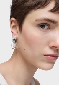 Pendientes de plata en forma de aro con tres círculos entrelazados, reflejando la luz. Vista en primer plano de la oreja de un modelo, con cabello corto y tono de piel natural.