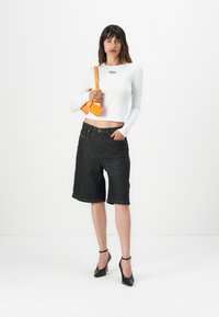 Bermudas en denim noir avec une coupe ample, associées à un crop top blanc à manches longues avec un petit accent métallique et un sac à main orange.