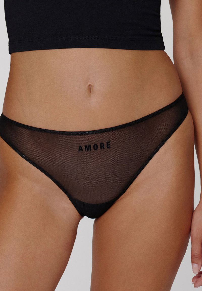 String transparent noir avec le texte "AMORE", une ceinture élastique lisse, et une couverture minimale en matériau en maille transparente.