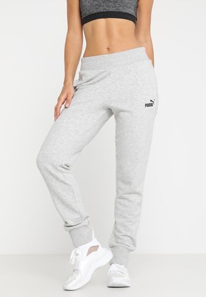 Vrouw die lichtgrijze Puma joggingbroek, witte sportschoenen en een donkergrijze sportbeha draagt, staand tegen een witte achtergrond.