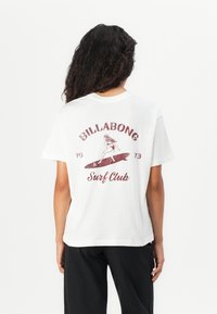 Persona con lunghi capelli ricci che indossa una maglietta bianca del Billabong Surf Club con un grafica vintage di un surfista e pantaloni neri, in piedi di spalle.