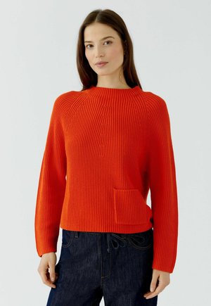 Vrouw met lang bruin haar die een feloranje gebreide trui draagt met een klein voorzakje en donkerblauwe spijkerbroek.