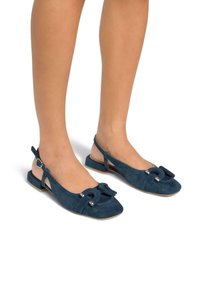 Ballerines en daim bleu marine à talon plat, avec un détail de nœud à l'avant et des accents métalliques sur le nœud. Présente un design à dos ouvert.
