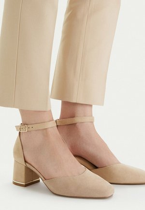 Zapatos de tacón grueso de ante beige con correas en el tobillo, usados con pantalones cortos a juego sobre fondo claro.