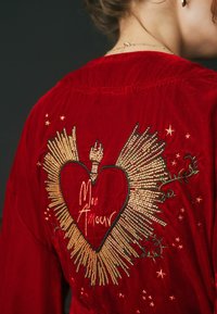 Červený sametový sako s veľkým vyšitým srdcom, zlatými akcentmi a hviezdičkovými vzormi. Na srdci je nápis "Mon Amour."