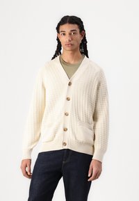 Cream knitted cardigan with a textured pattern, v-neck, six buttons, and two front pockets. Paired with a green shirt and dark jeans.