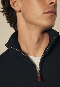 Donker marineblauwe zippered pullover met een getextureerd breiontwerp. Kenmerken zijn een hoge kraag, zilveren rits en een leren accent bij de trekker.