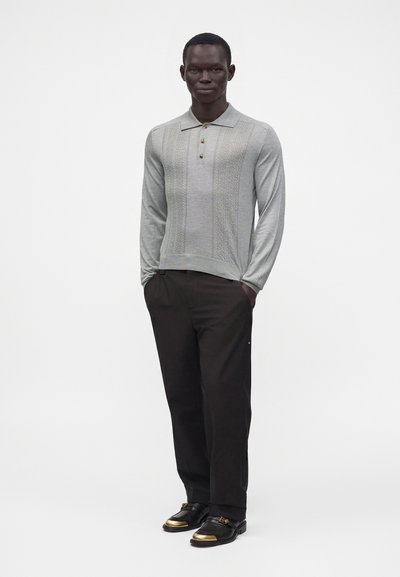 Homme debout les mains dans les poches, portant un polo en maille à manches longues gris clair, un pantalon noir et des chaussures noires avec des embouts dorés.