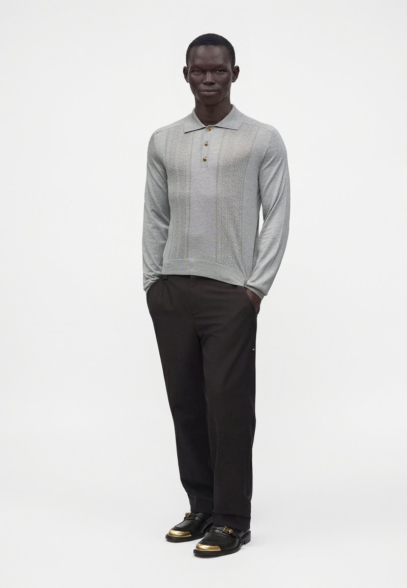 Homme debout les mains dans les poches, portant un polo en maille à manches longues gris clair, un pantalon noir et des chaussures noires avec des embouts dorés.