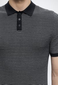 Homme portant un polo à motifs noir et blanc avec trois boutons noirs et manches courtes sur un fond uni.