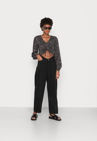 Blusa preta cropped com um padrão texturizado, mangas longas e laços na cintura; combinada com calças largas de cintura alta pretas e sandálias.