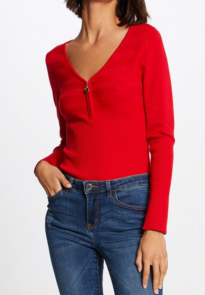 Femme portant un pull rouge à manches longues col en V avec fermeture éclair sur le devant et un jean bleu, main dans la poche.
