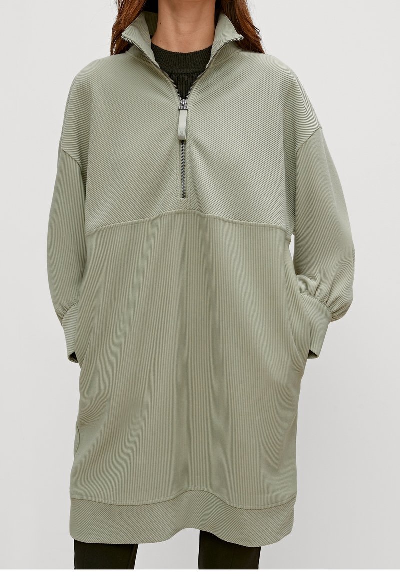 Femme vêtue d'une robe-pull côtelée verte claire oversize avec fermeture éclair à l'avant et poches latérales, mains dans les poches.