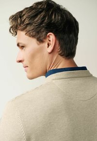 Polo tricot beige avec un col bleu. Présente une finition texturée et des accents côtelés au niveau du col, mettant en avant un style décontracté classique.