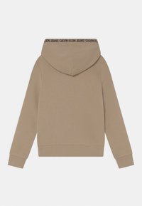 Beige mikina s kapucí, dlouhými rukávy a žebrovanými manžetami, na kterých je vytisknutý text "Calvin Klein Jeans" podél okraje kapuce.