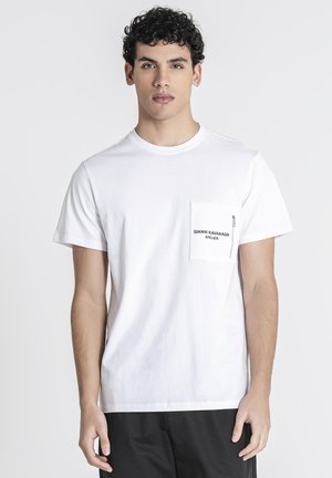 Mand med mørkt krøllet hår iført en hvid T-shirt med lynlåslomme på brystet og sorte bukser, stående mod en ensfarvet baggrund.