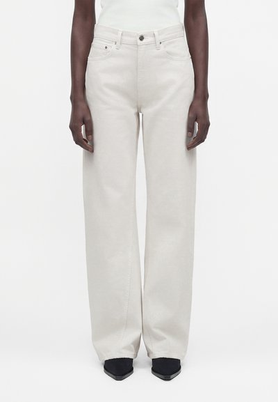 Personne portant un pantalon taille haute, jambes larges, couleur blanc cassé, avec boutons et poches à l'avant, associé à des chaussures noires pointues sur fond blanc.