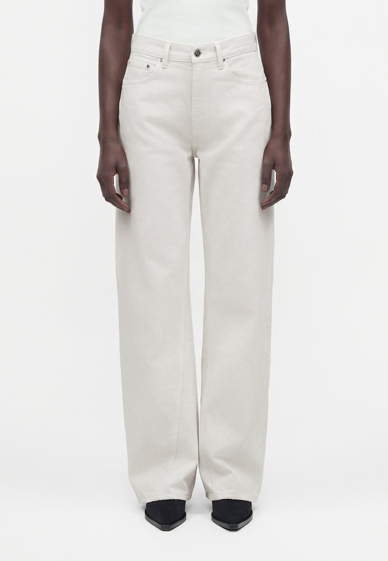 Personne portant un pantalon taille haute, jambes larges, couleur blanc cassé, avec boutons et poches à l'avant, associé à des chaussures noires pointues sur fond blanc.