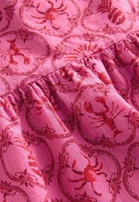 Tissu rose présentant un motif répété de crabes rouges entourés de motifs floraux, mettant en avant une texture douce et un détail froncé le long de la couture.
