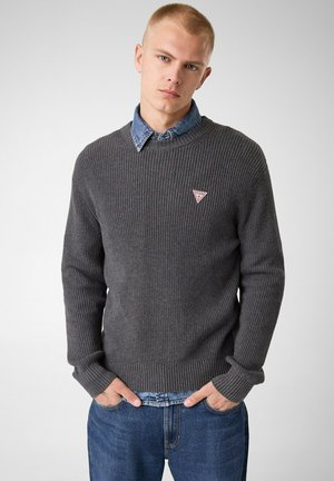 Ung mand med kort blondt hår iført en grå strikket sweater over en denimskjorte og blå jeans, hænderne i lommerne, neutralt baggrund.