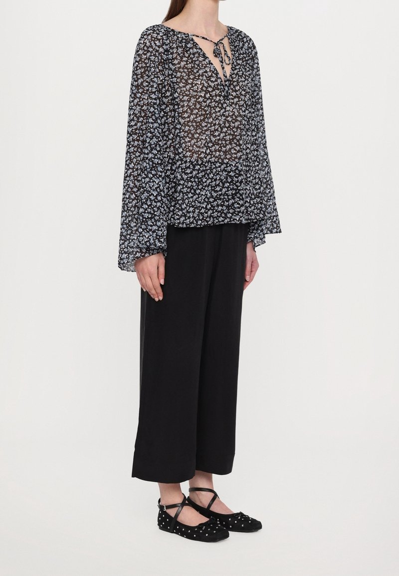 Femme portant un chemisier noir transparent à motif floral avec des manches cloches, un pantalon noir à jambes larges et des ballerines noires cloutées avec des brides à la cheville.