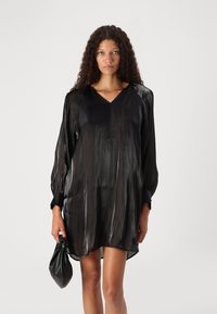 Rochie din mătase neagră, cu decolteu în V, mâneci lungi și o strălucire subtilă. Prezintă o croială confortabilă și slituri laterale, combinată cu o geantă mică neagră.