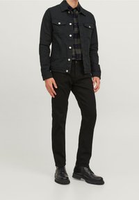 Giacca di jeans nera con due tasche sul petto, abbinata a pantaloni neri e stivali neri stringati. L'outfit presenta una camicia a quadri scuri sotto.