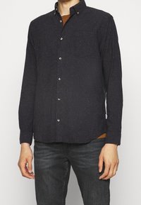 Marinblå knäppskjorta i texturerad tyg med flerfärgade fläckar, bröstficka och krage med knappar. Bärs över en brun topp.