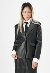 Svart lærblazer med strukturerte skuldre, to frontlommer og en enkelt knapplukking, sammen med en hvit skjorte og svart slips.