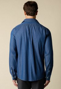 Mężczyzna w niebieskiej koszuli dżinsowej z długim rękawem i guzikami na mankietach, widziany z tyłu na tle gładkiego beżowego tła.