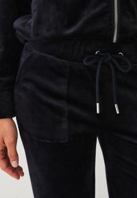 Survêtement en velours bleu marine avec ceinture élastique, fermeture à cordon de serrage et poche latérale. Texture lisse avec une finition douce. Détails en métal argenté.