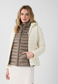 Gewatteerde jas in crème en lichtbruin, met een ritssluiting aan de voorkant, een opstaande kraag en een horizontale doorgestikte structuur. Twee zijzakken zichtbaar.