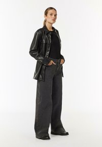 Schwarze Kunstlederjacke, schwarzes enges Oberteil, weite schwarze Jeans mit silberfarbigen Nietenakzenten, schwarze Stiefel mit Gummisohle, neutraler Hintergrund.