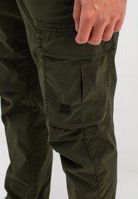 Olijfgroene cargobroek met meerdere zakken, een gestructureerde stof en een subtiele logo-applicatie op de linker bovenbeen. Verstelbare tailleband.