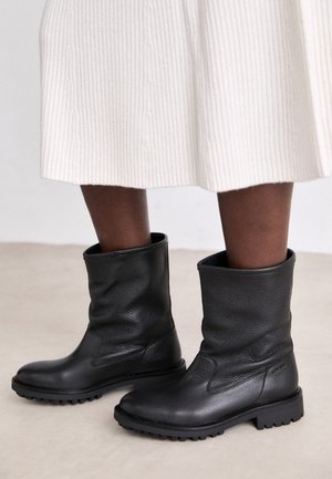 Personne portant des bottes en cuir noir mi-mollet avec des semelles texturées et une jupe blanche en maille côtelée, debout sur un sol clair.