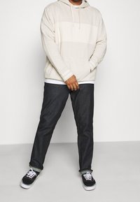 Mann in beiger Kapuzenjacke, weißem Unterhemd, dunklen hochgekrempelten Jeans und schwarzen Turnschuhen, stehend vor einem einfachen weißen Hintergrund.