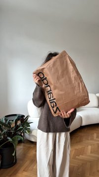 Henkilö, joka pitää suurta ruskeaa paperikassia, jossa on "Zalando"-teksti, seisoo olohuoneessa, jossa on valkoinen sohva, puulattia ja ruukkukasvi.