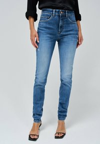 Jeans de denim ajustados en un lavado azul medio con decoloración, detalles desgastados, tiro alto, diseño de cinco bolsillos y cierre de botón.