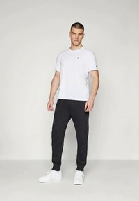 Valkoinen V-kaula-aukkoinen t-paita, jossa on pieni logo rinnassa, yhdistetty mustiin verkkareihin. Mallilla on päällään valkoiset urheilukengät, mikä luo rentoa asustusta.