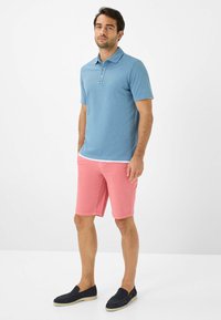Polo shirt azzurro chiaro con colletto, abbinato a pantaloni corti rosa. I pantaloni corti hanno una texture liscia e il modello indossa scarpe slip-on scure.
