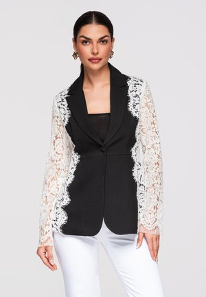 Žena v černém saku s bílými krajkovými rukávy a bílých kalhotách, stojící před světle šedým pozadím.