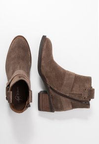 Botas de ante marrón hasta el tobillo con un tacón apilado, cremallera lateral y una correa con un detalle de hebilla. Textura suave y diseño de punta redonda.