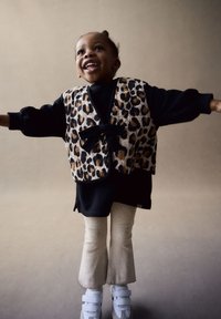 Chaleco con estampado de leopardo y lazo, camisa negra de manga larga, pantalones acampanados color crema y zapatillas blancas. Texturas suaves y colores contrastantes.